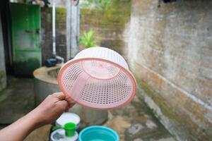 A hand holds a pink plastic strainer, with a blurred background of green plants and stone. photo