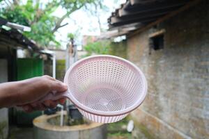 A pink plastic strainer held in a hand, a well in the background. photo