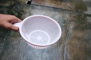 A hand holds a white plastic strainer with a grid pattern against a blurred concrete background. photo
