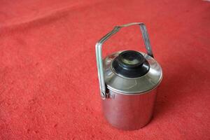 A silver metal container with a black lid and handle sits on a red surface. photo