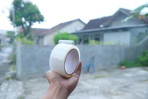 A hand holds a roll of clear packing tape with a blurred background of a home and greenery. photo