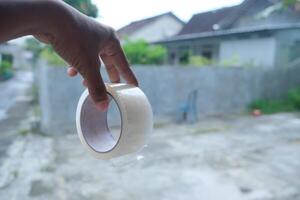 A hand holds a roll of clear packing tape in front of a blurred background. photo