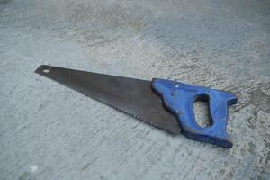 A blue-handled hand saw lies on a rough concrete surface. photo