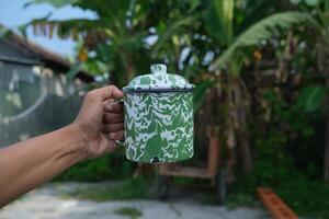 A hand holding a green and white enamel mug with a blurred background of green foliage. photo