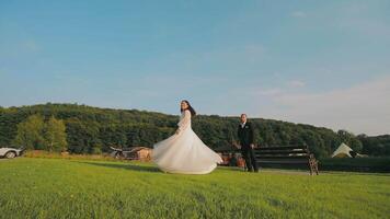 Bride Twirling in a White Gown Outdoors with Groom Watching. A bride twirls joyfully in her flowing white gown on a grassy field as the groom stands watching nearby. video
