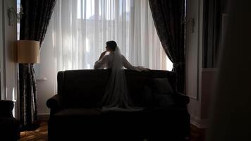 Bride reflecting by the window in a softly lit room. A bride stands thoughtfully by the window in a dimly lit room, with sheer curtains softly filtering the light. video