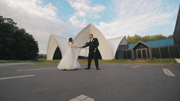 Bride and Groom Dancing Outdoors by Modern Architecture. A bride and groom dressed in formal wedding attire are dancing outdoors in front of a modern architectural building with a unique design. video