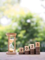 Hourglass, Wooden blocks with the number 2025 on stack of coins. The concept of saving money for manage time to success business in year 2025 photo