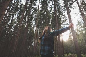 Happy woman in a plaid shirt with outstretched arms enjoying the serenity of a dense forest, surrounded by tall trees, feeling freedom and connection with nature photo