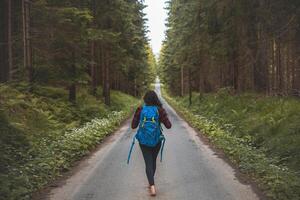 Back view of a woman with a blue backpack walking barefoot down a peaceful forest road, surrounded by lush greenery and tall trees, symbolizing adventure and tranquility in nature photo