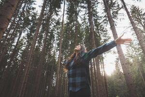 Happy woman in a plaid shirt with outstretched arms enjoying the serenity of a dense forest, surrounded by tall trees, feeling freedom and connection with nature photo