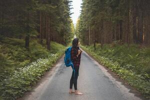 Back view of a woman with a blue backpack walking barefoot down a peaceful forest road, surrounded by lush greenery and tall trees, symbolizing adventure and tranquility in nature photo