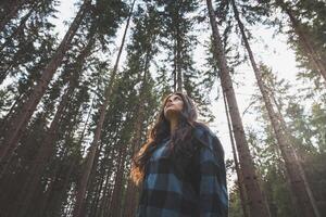 Happy woman in a plaid shirt with outstretched arms enjoying the serenity of a dense forest, surrounded by tall trees, feeling freedom and connection with nature photo