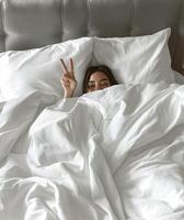 A person relaxing in bed surrounded by white linens with a peace sign gesture during morning photo