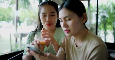 Footage of handheld shot, Two siblings happily sit at a table, drinking cold drinks while playing social media together on smartphone at a coffee shop. Family enjoying happy time together. video