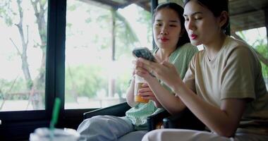 Handheld selective focus shot, Two siblings happily sit at a table, drinking cold drinks while playing social media together on smartphone at a coffee shop. Family enjoying happy time together. video