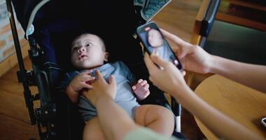 Handheld selective focus shot, Two women are having fun playing with little boy in a baby stroller and recording a cute on mobile phone in a coffee shop. Family enjoying happy time together video