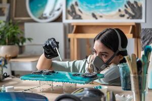 Female artisan in respirator drips liquid paint onto epoxy resin artwork to create waves in workshop photo