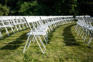 Folding chairs standing in row on green grass, casting shadow on ground. Outdoor party preparation photo
