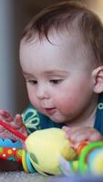 Bright toy in the hands of a baby lying on the carpet. Focused toddler boy playing with a toy indoors. Blurred backdrop. Vertical video