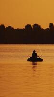 barco com uma pescador dentro a pôr do sol luz flutua lentamente em a lago. silhueta do uma homem sentado dentro uma barco e pescaria em a laranja fundo do lindo pôr do sol dentro a rio. vertical video