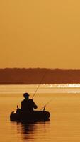 Silhouette of a man in hat fishing from the boat on the evening river background. Beautiful sunset and a fisherman catching fish from the inflatable boat in the open water. Vertical video