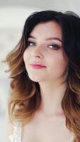 Portrait of a beautiful young bride with wavy hairdo smiling into camera. Glamour model with stylish makeup is sitting on bed and posing in the studio. Vertical video