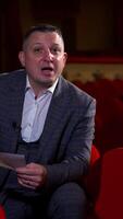 Man actor gives interview while sitting on red velvet chair in classic theater. Empty theter before evening performance. Vertical video