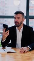 Male businessman sitting at the table is going to make a call. Man takes a paper notebook and pen to make some notes. Office backdrop. Vertical video
