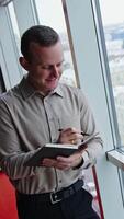 Caucasian man standing at the window and taking notes into notebook. Thoughtful male office worker at the backdrop of cityscape. Vertical video