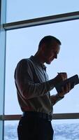 Tall slim man stands against the panoramic window. Male taking notes, looks into window and throws away one page. City view at backdrop. Vertical video