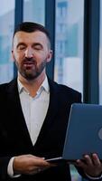 Smiling good-looking bearded businessman in suit stands near the window holding laptop. Man talks showing something with his hands. Vertical video