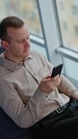 Middle-aged man sitting in the bean bag chair and holding a phone. Man is glad about message and writes it back. High angle view. Vertical video