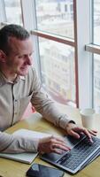 Happy smiling confident man working on laptop. Notebook and cup in front of him on the table. High angle view. Cityscape at backdrop. Vertical video