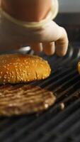 Side view of the delicious burger cutlet and two buns flipped on the grill with hand in latex gloves. Selective focus. Vertical video