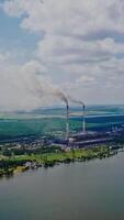 Aerial view of plant on the bank of the river in summer. View from above on natural background of beautiful river and factory with smoke from pipes. Slow motion. Camera moves up. Vertical video