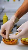 Hands in latex gloves put pickles on top of fried bun. Making burger on white table. Fast food concept . Vertical video
