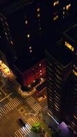 Dark multi-storied buildings with lit windows at night. Drone descending over the well-lit roads with busy traffic. Vertical video