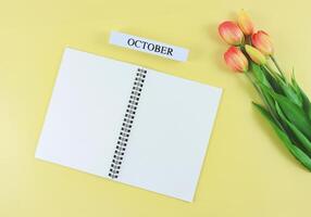 flat lay of opened blank pages notebook or diary, wooden calendar October with tulips bouquet on yellow background with copy space. photo