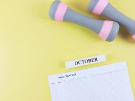 flat lay of habit tracker book, wooden calendar October, gray pink dumbbells, on yellow background with copy space. Self development and active lifestyle concept. photo