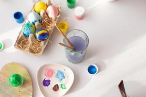 On a white table, in daylight, there are brushes in a glass of water, multi-colored eggs in a tray photo