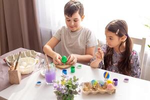 A happy boy and little girl sits at a white table near the window, coloring eggs photo