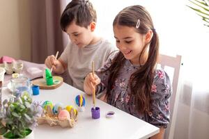 A smiling little girl and boy sits at a white table near the window, paints eggs photo