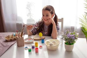 Happy little girl sits at a white table by the window, holds a brush, a coloring egg photo