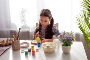 A adorable little girl sits at a white table by the window, paints eggs photo