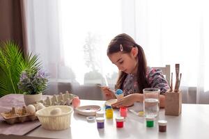 A cute little girl sits at a white table by the window, paints eggs with a brush and paint in jars. photo