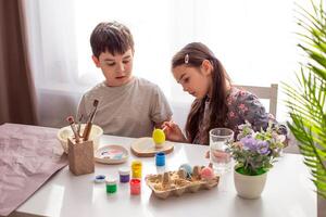 A brunette little girl and boy sits at a white table near the window, paints eggs photo