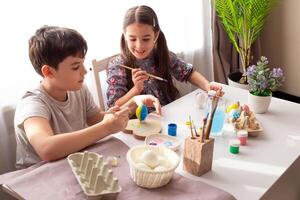 Smiling little girl and boy are sitting at a white table, coloring eggs for the Easter holiday photo