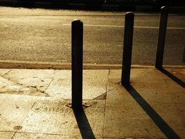 A street with three metal poles on the sidewalk. The shadows of the poles are cast on the ground photo