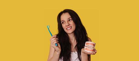 A young Caucasian brunette woman with a toothbrush and a jaw model on a yellow background. A panoramic banner with a place for your text. photo